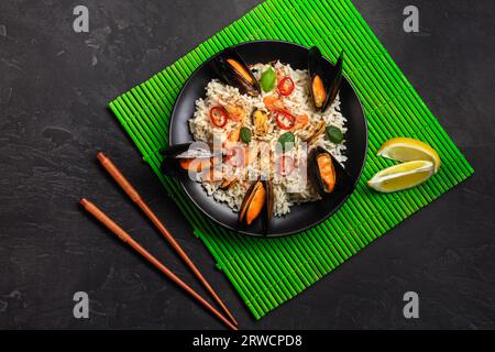 Riso fritto con frutti di mare cozze, gamberi e basilico in una piastra nera con bacchette in bambù verde mat e tavolo di pietra. Vista dall'alto. Foto Stock