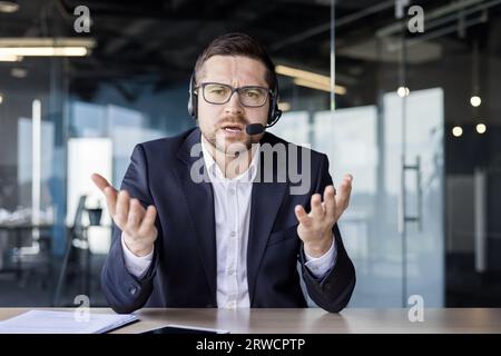 Ritratto di un giovane stanco e turbato in un auricolare seduto in ufficio di fronte alla fotocamera e infelice parlare durante una videochiamata. Si alza e si muove con le mani. Foto Stock
