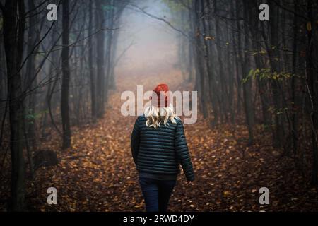 Donna solitaria che cammina su un sentiero nella foresta nebbiosa e scura. Atmosfera spettrale nei boschi autunnali Foto Stock