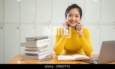 Una giovane studentessa asiatica sorride davanti alla macchina fotografica e siede a un tavolo con il suo portatile e una pila di libri sulla taia Foto Stock