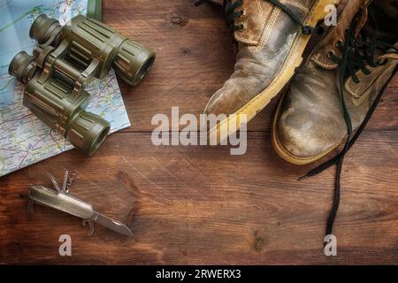 Vecchi scarponi da trekking in pelle, binocoli, mappa e coltello tascabile su legno rustico scuro, concetto per viaggi, escursioni e vacanze avventurose, vista dall'alto Foto Stock