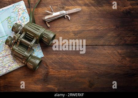 Binocolo, mappa e coltello tascabile su uno sfondo rustico scuro in legno, concetto di viaggio per trekking, escursioni e vacanze avventurose, vista dall'alto Foto Stock