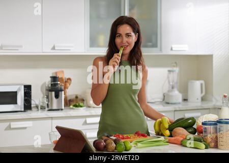 Ritratto di una giovane donna in piedi al bancone della cucina, mordere il gambo di sedano e guardare la macchina fotografica Foto Stock