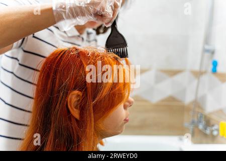 Il lavoro di un maestro della colorazione dei capelli. Il processo di tintura dei capelli rossi. Le mani del maestro indossate con guanti protettivi applicano una spazzola sulle radici dei capelli Foto Stock