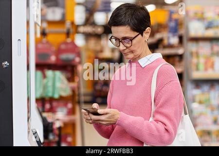 Ritratto di una donna adulta che usa lo smartphone al momento del check-out in un supermercato Foto Stock