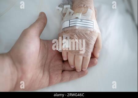 Il genitore tiene la mano del bambino con la vista ravvicinata del gocciolamento IV sullo sfondo del letto Foto Stock