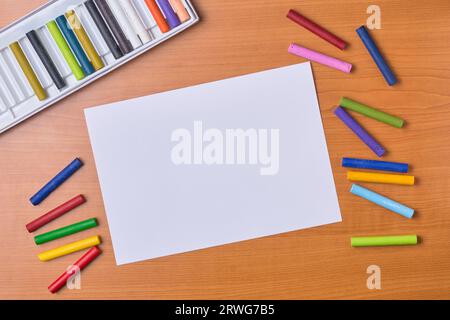 pastelli ad olio e carta bianca vuota o bianca sulla scrivania, pastelli sparsi e set box sullo sfondo, presi direttamente dall'alto Foto Stock