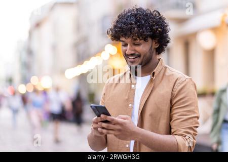 Giovane indiano sorridente che sta fuori e usa il telefono, invia SMS, chatta, chiama. Foto Stock