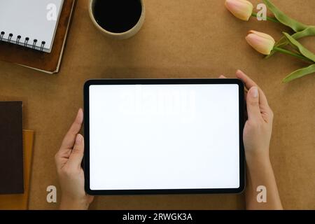 La mano di una donna regge il modello di tablet digitale con schermo vuoto, modello didattico e di concetto applicativo, sfondo marrone chiaro, vista dall'alto e spazio per la copia Foto Stock