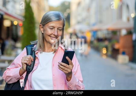 Turista matura sorridente che digita un messaggio al telefono, donna in viaggio pensionato dai capelli grigi che usa l'app per smartphone mentre cammina in città sorridente sorridente. Foto Stock