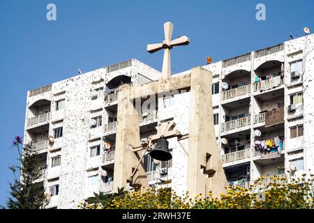 Campanile con una croce sopra una chiesa di fronte a edifici residenziali coperti da buchi di proiettile a Tripoli, Libano Foto Stock