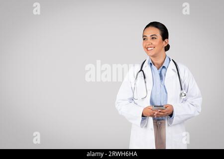 Felice medico brasiliano in cappotto bianco che usa il cellulare e guarda da parte lo spazio libero, dando consulenza online Foto Stock