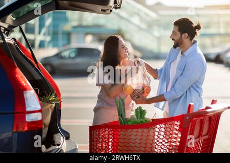 Felice coppia latina in piedi nell'area parcheggio e caricando i sacchetti di carta dopo aver fatto acquisti nel bagagliaio aperto Foto Stock