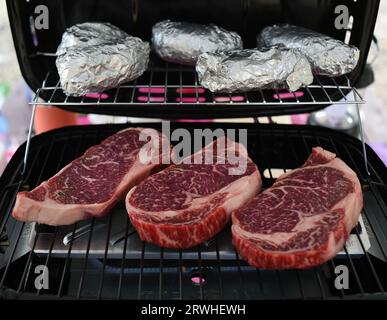 Le bistecche di manzo Wagyu ben marmorizzate cucinano su un barbecue insieme a patate ricoperte di foglio di alluminio Foto Stock