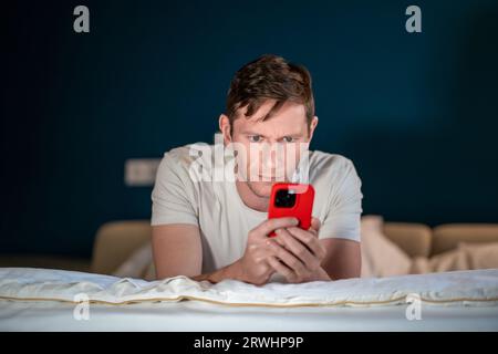 L'uomo usa lo smartphone sdraiato sul letto. Acquisti online su Internet risoluzione di problemi aziendali conduce chat Foto Stock