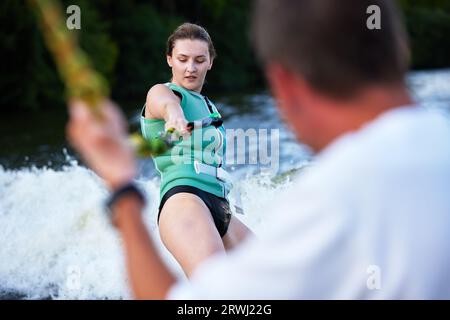 Una giovane ragazza sportiva con un giubbotto di salvataggio verde cavalca un wakeboard su un fiume o un lago vicino alla città. La felice atleta scivola sull'onda seguendo la Foto Stock