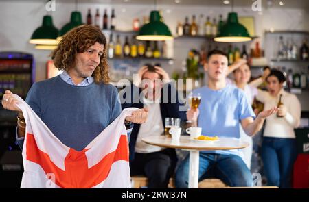 Compagnia di giovani tifosi adulti con bandiera nazionale d'Inghilterra sconvolta per la squadra preferita che perde partita in bar Foto Stock