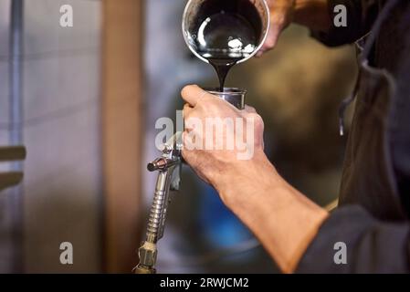 Mani di una giovane persona sconosciuta che versa vernice in un serbatoio di una pistola a spruzzo nella sua officina. Vista ravvicinata. Foto Stock