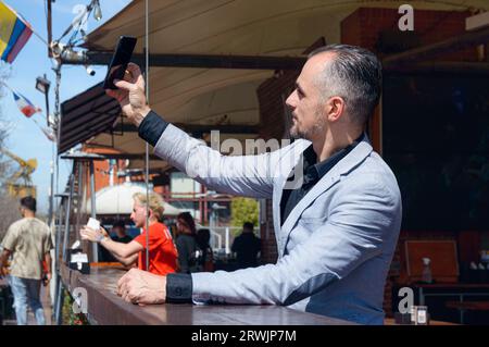 Uomo d'affari caucasico adulto con elegante giacca in tuta che scatta selfie con il telefono seduto fuori dal ristorante, concetto tecnologico, spazio fotocopie. Foto Stock