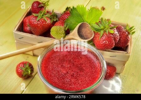 Marmellata di fragole fatta in casa in un barattolo accanto a un cestino di fragole fresche, cucchiaio da cucina e zucchero Foto Stock