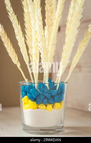 Orecchie di grano in vaso blu e giallo. Piante essiccate in vaso colorato ucraino. Grano bianco su ripiano di legno bianco. Elegante arredamento ecologico. Bouquet di cereali. Foto Stock