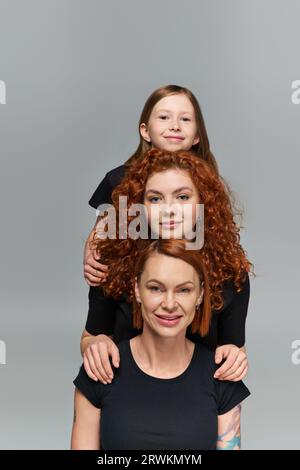 concetto di generazione femminile, famiglia rossa positiva in abbigliamento coordinato abbracciato su sfondo grigio Foto Stock