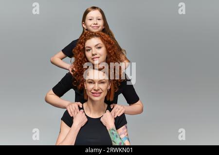 concetto di generazione femminile, famiglia happy redhead in abbigliamento coordinato abbracciato su sfondo grigio Foto Stock