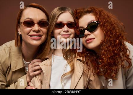 concetto di cambiamento di generazione familiare, donne felici rosse e ragazza con occhiali da sole su sfondo marrone Foto Stock