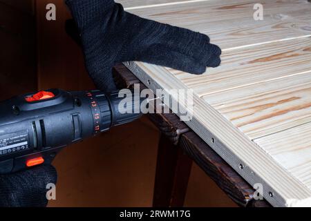 Un falegname trapana un foro in una tavola di legno con un trapano elettrico. Primo piano delle mani di un caposquadra al lavoro. Ambiente di lavoro in una falegnameria Foto Stock