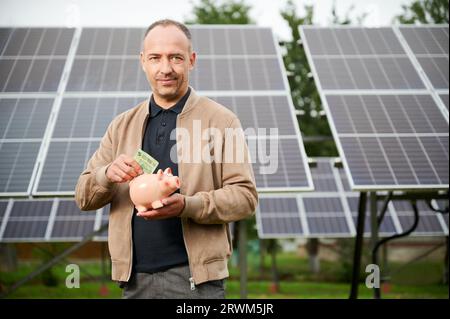 Uomo di fiducia che mette i soldi guadagnati alla banca del piggy. Il profitto di risparmio dell'adulto maschio ha ottenuto investendo nell'energia verde. Uomo in abiti casual tenendo in mano la scatola del denaro e mettere euro in esso. Foto Stock