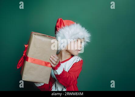 felice sorridente ragazzo caucasico con maglietta e cappello di babbo natale che regge una scatola con regalo di natale avvolta in carta artigianale. Bambino isolato su sfondo verde Foto Stock