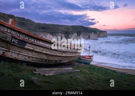 Barche da pesca sopra una spiaggia sulla costa dello Yorkshire a Flamborough e in una baia circondata da alte scogliere. Onde intorno alle rocce e cielo in tarda serata. Foto Stock