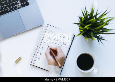 aprite il blocco note e le risoluzioni di capodanno per la scrittura a mano delle donne. Fiore, computer portatile e tazza di caffè sullo sfondo. Foto Stock