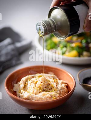 l'olio d'oliva dorato viene versato delicatamente da una bottiglia a un recipiente riempito con hummus cremoso. Sullo sfondo si aggiunge un piatto di verdure fresche Foto Stock