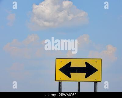 Un segnale stradale isolato giallo e nero a doppia freccia con un cielo blu nuvoloso sullo sfondo Foto Stock