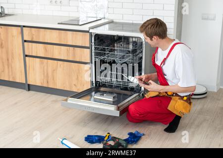 Il tecnico o il lavoratore in uniforme installa la lavastoviglie nei mobili della cucina. Il riparatore indossa tuta da lavoro riparando la manutenzione della lavastoviglie. Maestro Foto Stock