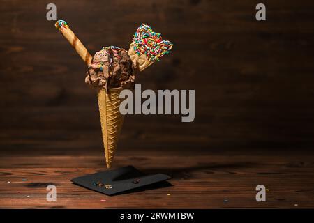 divertente concetto creativo di cono cialde volante con gelato coperto, spruzzi di sigari decorati e ventole versate con glassa di cioccolato su legno. Foto Stock