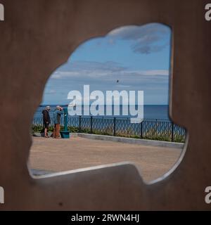 Vista attraverso la scultura del castello di sabbia di Richard Broderick sulla passeggiata centrale, due uomini usano il telescopio a Whitley Bay con il faro di St Mary Foto Stock