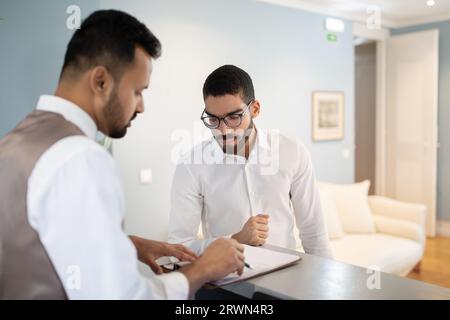 Un uomo d'affari arabo che effettua il check-in all'hotel, firmando i documenti con la receptionist Foto Stock