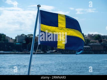 Bandiera nazionale della Svezia, sfocatura degli edifici di Stoccolma sul lungomare e sfondo blu del cielo, giorno di sole, Foto Stock