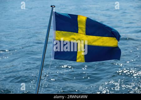 Bandiera nazionale della Svezia che sventola sull'asta di bandiera della barca, fondo di acqua di mare sfocata, giorno di sole, Foto Stock