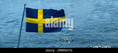 Bandiera nazionale della Svezia che sventola sull'asta di bandiera della barca, fondo di acqua di mare sfocata, giorno di sole, Foto Stock