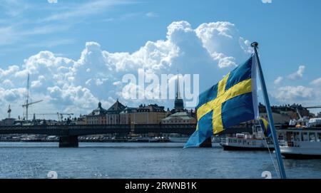 Bandiera nazionale della Svezia, sfocatura degli edifici di Stoccolma sul lungomare e sfondo blu del cielo, giorno di sole, Foto Stock