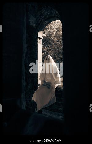 Una donna con un indumento bianco e spettrale con buchi per gli occhi seduta su una finestra in una chiesa abbandonata. L'immagine inquietante evoca temi di misticismo, occultismo, Foto Stock