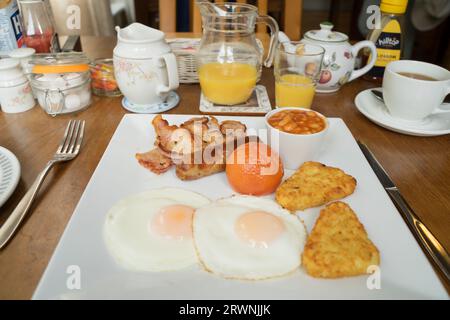Colazione all'inglese completa con un piatto, pancetta, salsiccia, uova fritte con contorno soleggiato, crocchette di patate, pomodoro arrosto, succo d'arancia in un barattolo, tazza da tè Inghilterra Regno Unito Foto Stock