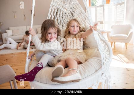 Due bambine cavalcano su un altalena di macrame a casa vicino all'albero di Natale. Le sorelle si divertono, ingannano in vacanza. Ragazze divertenti che hanno un hap Foto Stock