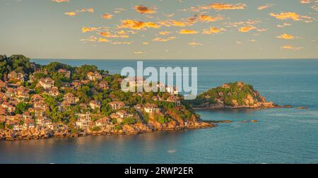 Bungalow fotografati dalla collina sulla costa di Ko Tao, Thailandia, nascosti tra palme e rocce e posti a gradini verso l'oceano. Nuvola d'arancio Foto Stock