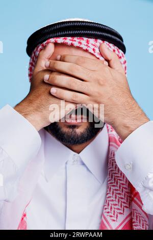 Uomo vestito con abiti islamici che chiude gli occhi con le mani mentre mostra che non si vedono segni malvagi tre scimmie sagge. Giovane musulmano che mostra il concetto di problema della vista in primo piano in studio Foto Stock