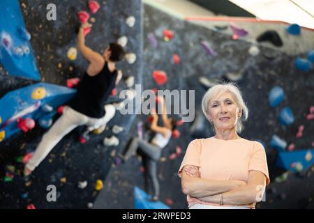 Donna anziana che indossa abbigliamento sportivo sorride alla macchina fotografica mentre si trova in piedi contro l'allenamento artificiale arrampicata parete di roccia nel parco avventura Foto Stock