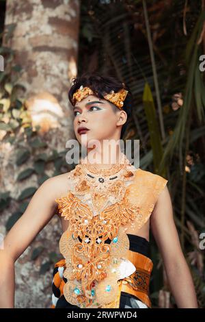 Un uomo balinese con un costume d'oro e un trucco sul viso si trova di fronte all'albero di cocco nella foresta Foto Stock